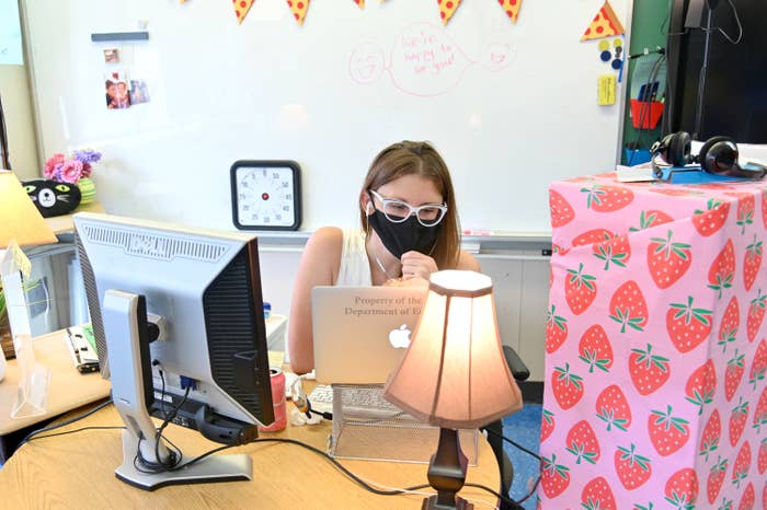 A teacher sits at her desk with her mask on, teaching on her laptop.