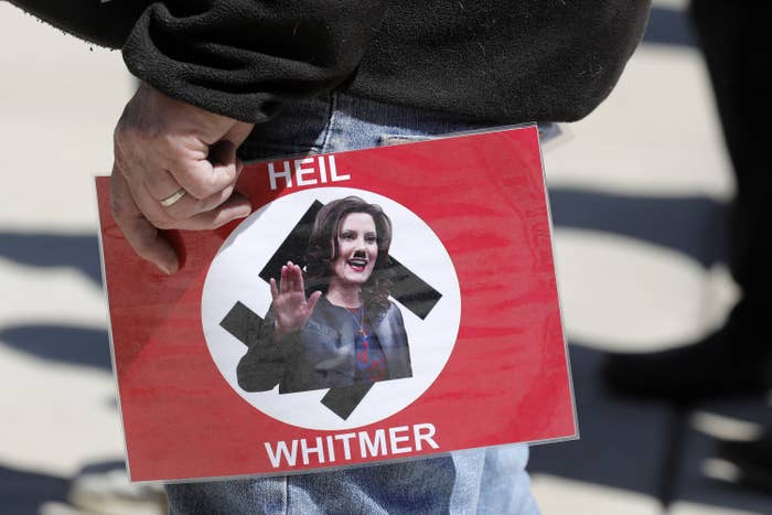 Someone holds a sign with the flag of Nazi Germany; over the swastika in the center, Gretchen Whitmer appears with a photoshoppted Hitler mustache, appearing to make a "Heil Hitler" salute; the sign reads "Heil Whitmer"