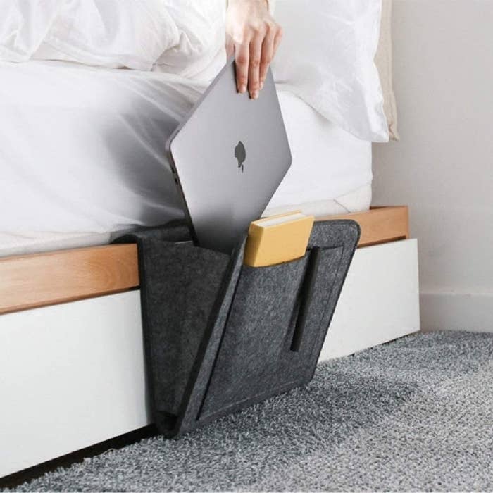 A close up of a person putting a laptop into the felt organizer tucked between a mattress and a bedframe