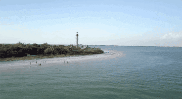 An aerial shot of a lighthouse by the beach with people swimming nearby.