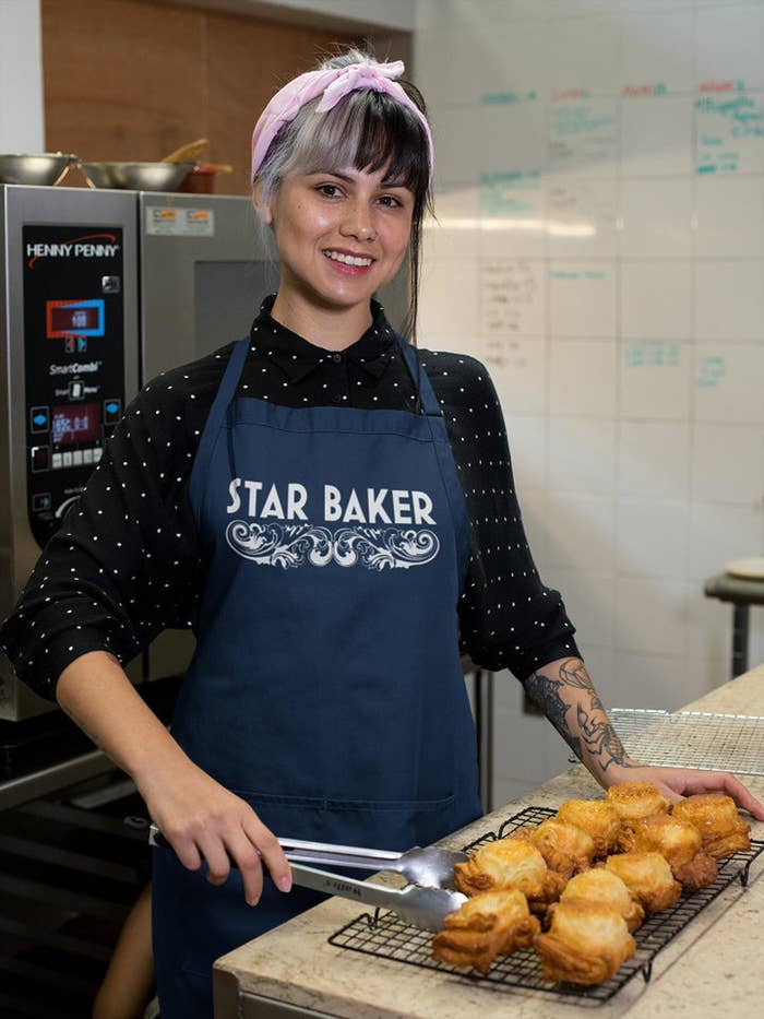 A model in the navy apron that says Star Baker with the GBBO scroll design underneath