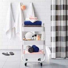 A white 3 tier cart on wheels filled with bathroom towels and accessories