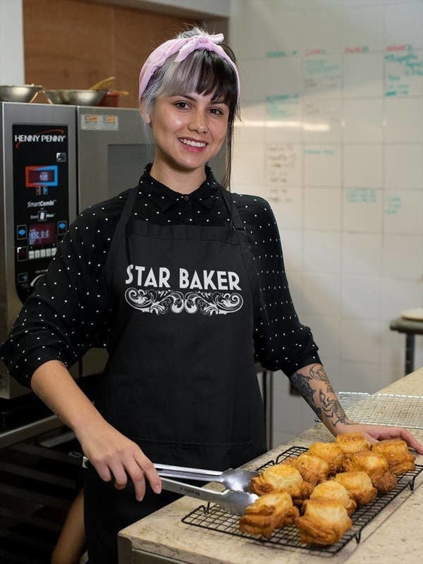 a model wearing a black apron with the words &quot;star baker&quot; on it in white and a decorative flourish below the words