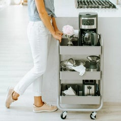 A white 3 tier cart on wheels filled with coffee and kitchen supplies