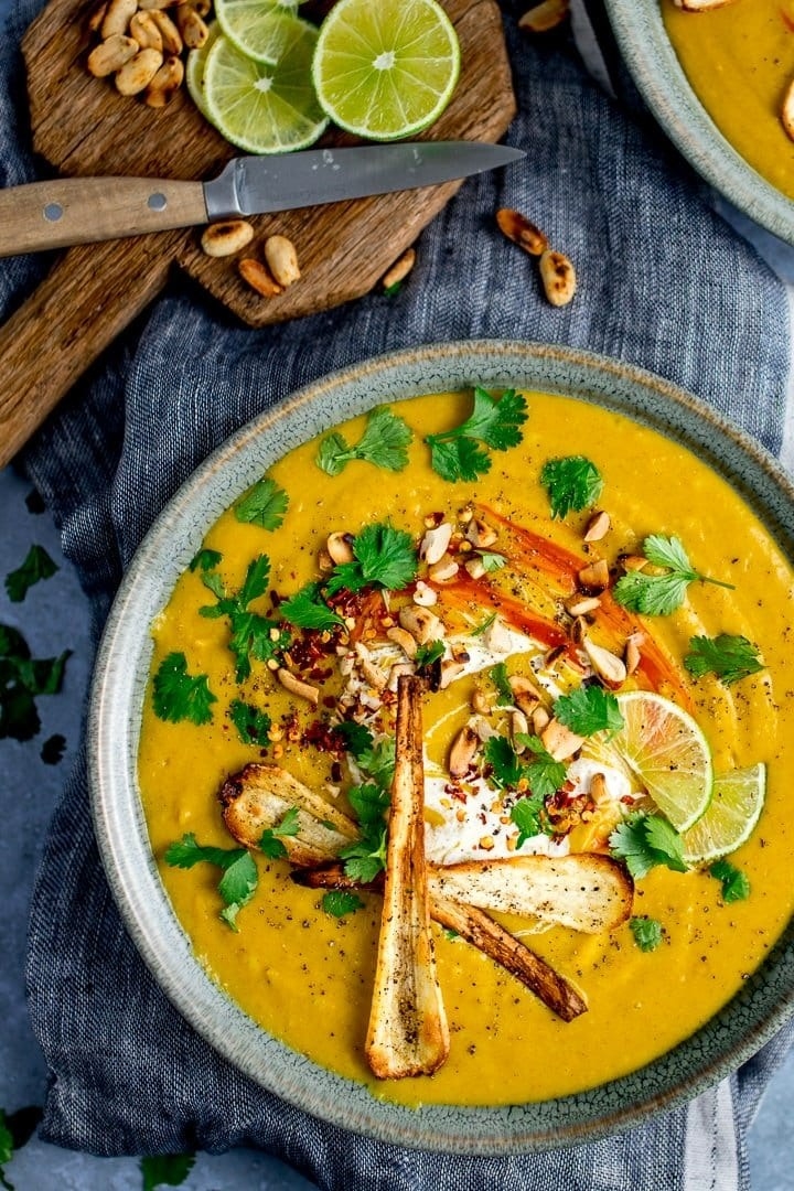 A bowl of creamy sweet potato soup with roasted parsnips on top, fresh herbs, nuts, and lime wedges.