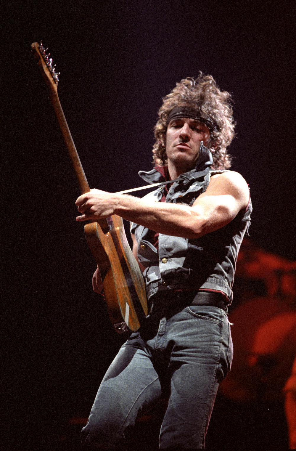 Bruce Springsteen performing onstage dressed in jeans, a denim vest, and a headband