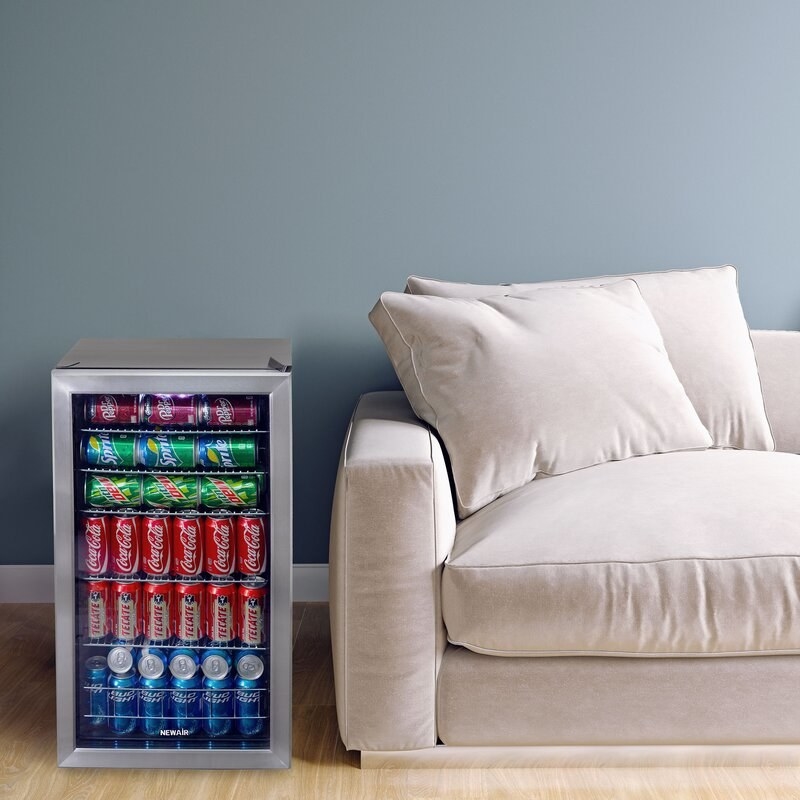 Gray drink fridge with silver and clear door beside a cream colored couch