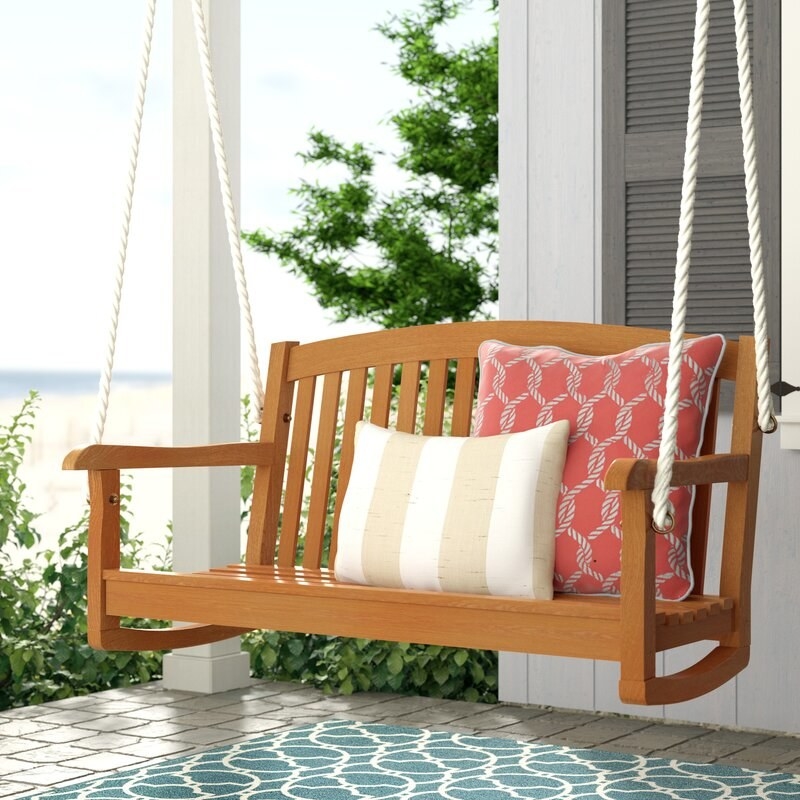 Brown porch swing with white ropes and displaying a white and tan striped pillow and red pillow with a white rope design