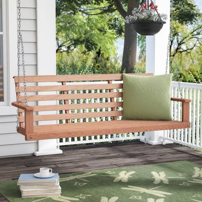 A wooden porch swing with metal chains set up on a patio with a green throw pillow and green rug