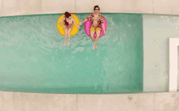 GIF showing top-down view of Andy Samberg and Cristin Milioti floating in a pool on donuts and clinking drinks for a cheers.