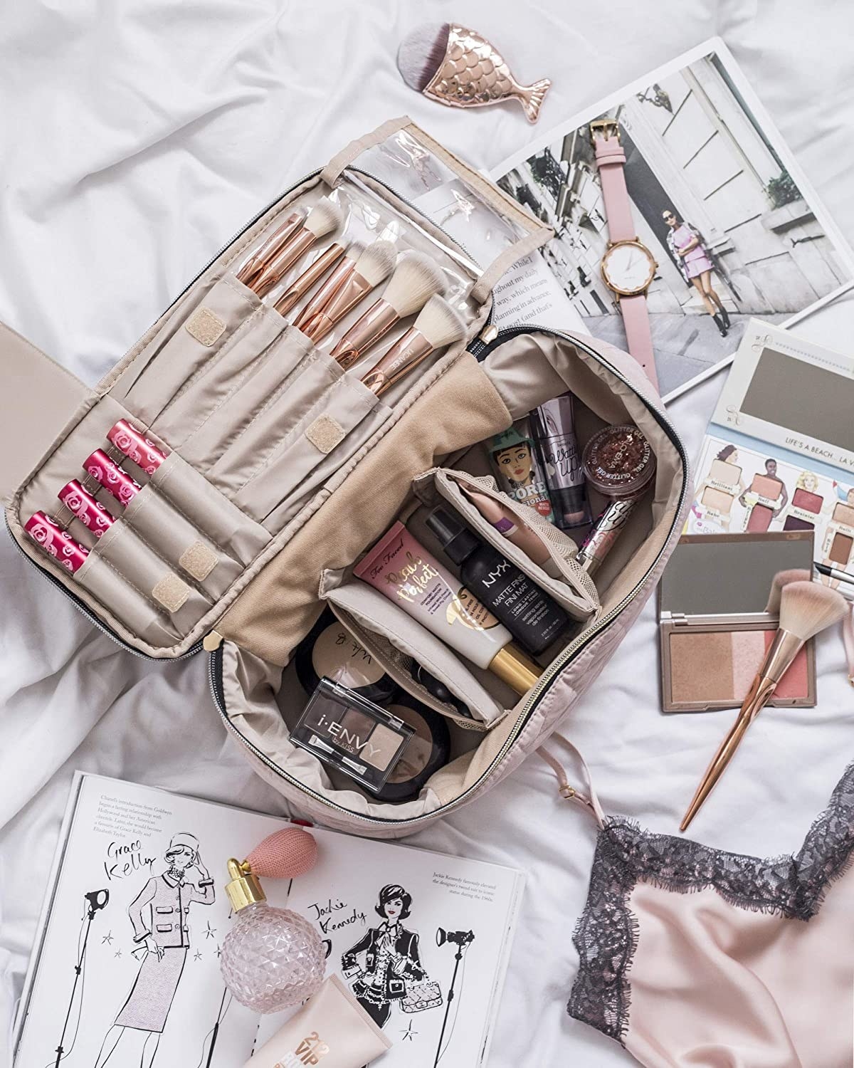 makeup bag filled with makeup surrounded by other products like books, a watch, perfume, and pajamas.
