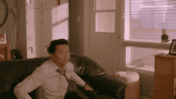 Jung (Simu Liu) falls onto his couch and crosses his legs on top of his coffee table as he clicks on the remote on Kim&#x27;s Convenience