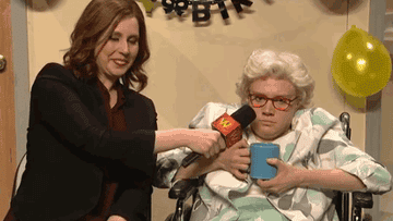 Vanessa Bayer holds a microphone up to Kate McKinnon, dresses as an old woman in a wheelchair, in a skit on SNL