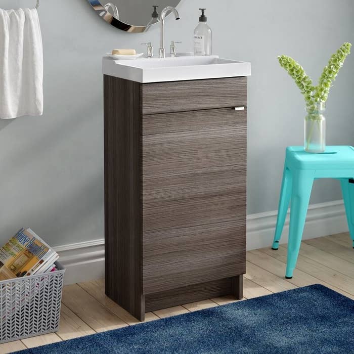 A very slim brown bathroom vanity with a small white sink next to a teal metal stool inside a bathroom