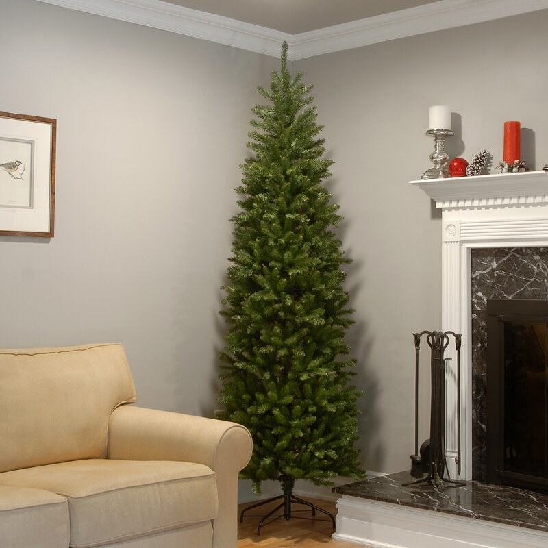 A slim green fake Christmas tree with no ornaments on it inside a family room, next to a couch and fireplace