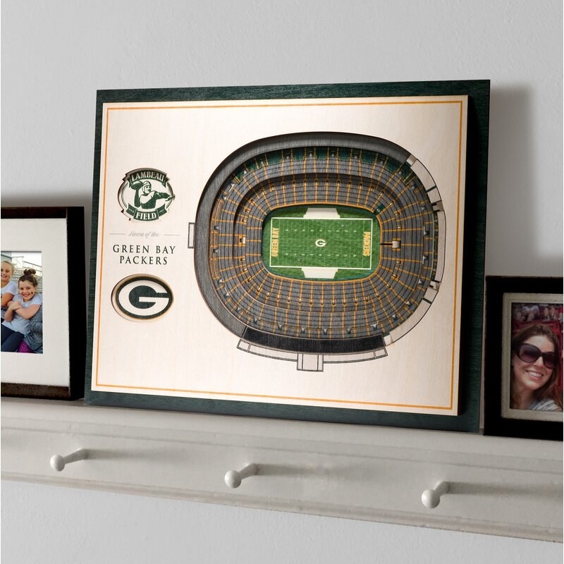 A framed recreation of the Green Bay Packers' NFL stadium on a white shelf next to framed family photos