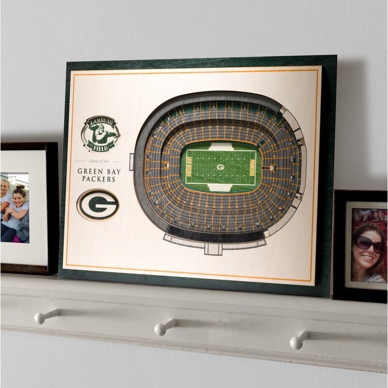 A framed recreation of the Green Bay Packers&#x27; NFL stadium on a white shelf next to framed family photos 