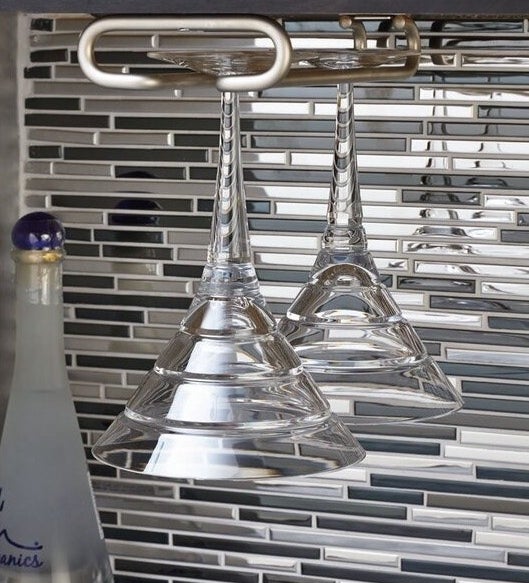 Two martini glasses hanging on a metal glass holder in front of a gray and silver backsplash 