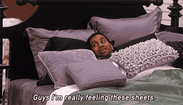 A GIF of a person lying in bed surrounded by pillow saying guys I&#x27;m really feeling these sheets very cozy