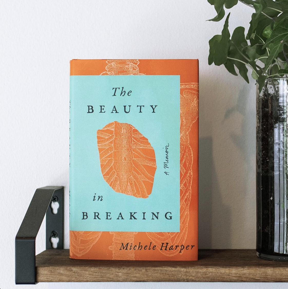 An upright copy of The Beauty in Breaking on a bookshelf with the cover showing an outline of a skeleton peeking through a teal rectangle with a hole in it
