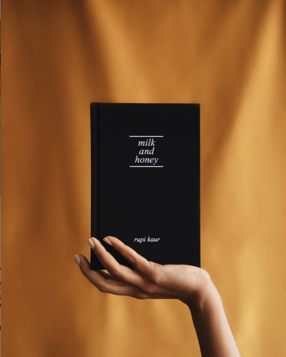hand holding the book Milk and Honey in an upright position in front of a yellow piece of fabric