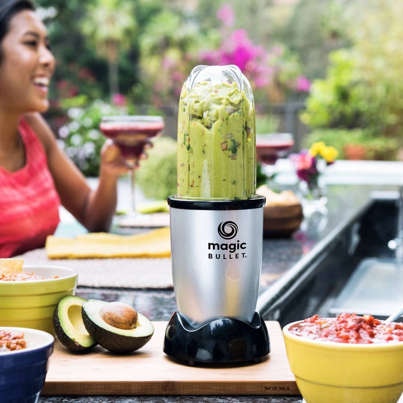 magic bullet with guacamole in it in the foreground and a woman drinking a margarita in the background