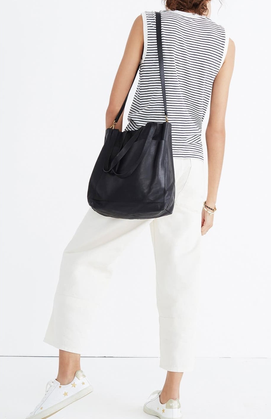 The black leather tote bag with shoulder straps over a model&#x27;s shoulder and a large pocket on the front with shorter straps