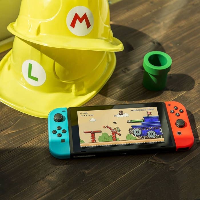 A blue and red Nintendo switch handheld on top of a wooden table with two yellow hats in the background, one with an M for Mario and another with an L for Luigi