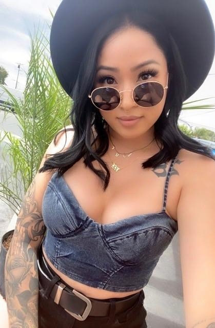Reviewer wears black round sunglasses while wearing a black fedora, a denim crop top, and belted black shorts