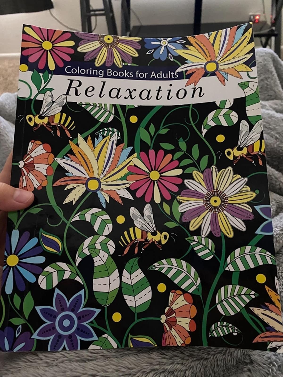 A reviewer holds up the cover of Coloring Books for Adults: Relaxation, depicting flowers and insects, that they purchased off of Amazon