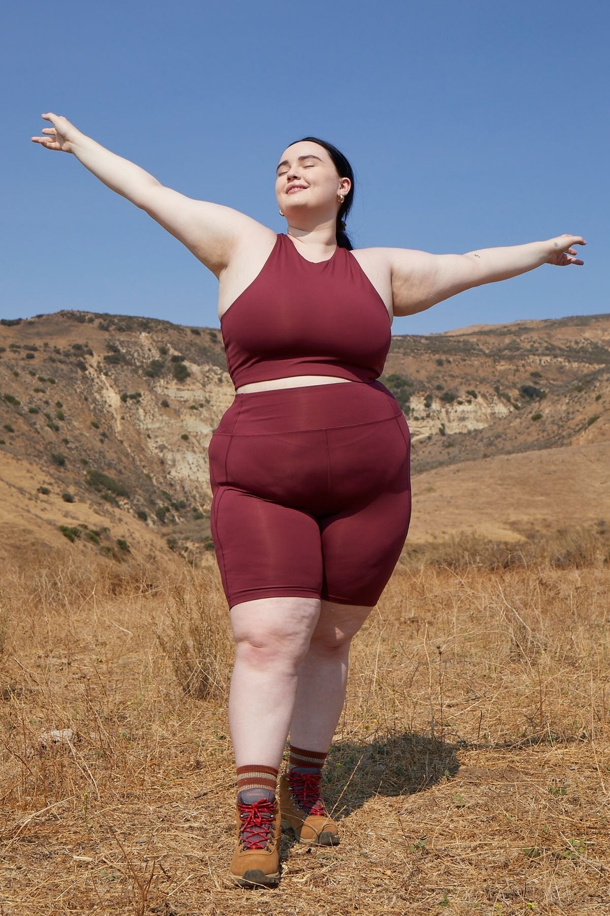 Model wears high-rise dark red bike shorts with a matching sports bra and brown hiking booties