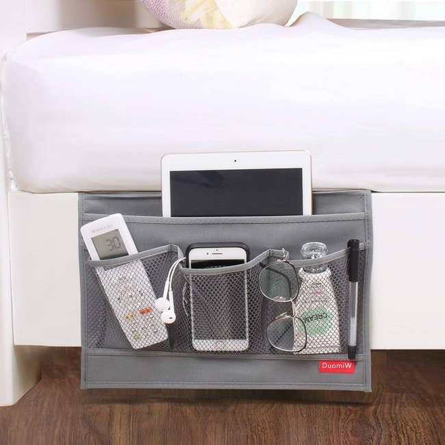 The grey bedside caddy holding a remote, iPad, cellphone, glasses, pen, lotion, and headphones