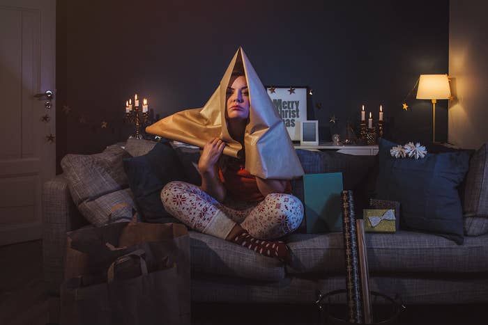 Woman sitting on couch with wrapping paper on her head surrounded by bags and holiday gifts 