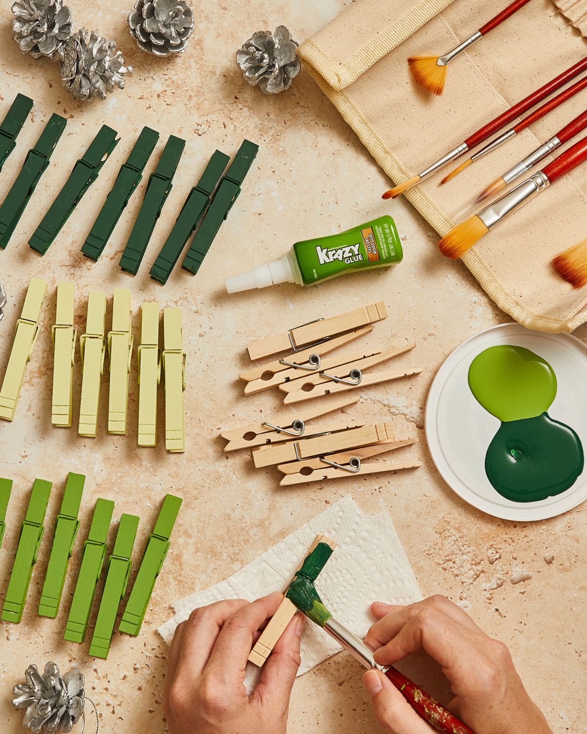 Clothespins, paintbrushes, and green paint laid out. Person is painting one of the clothespins green. Bottle of Krazy Glue off to the side.