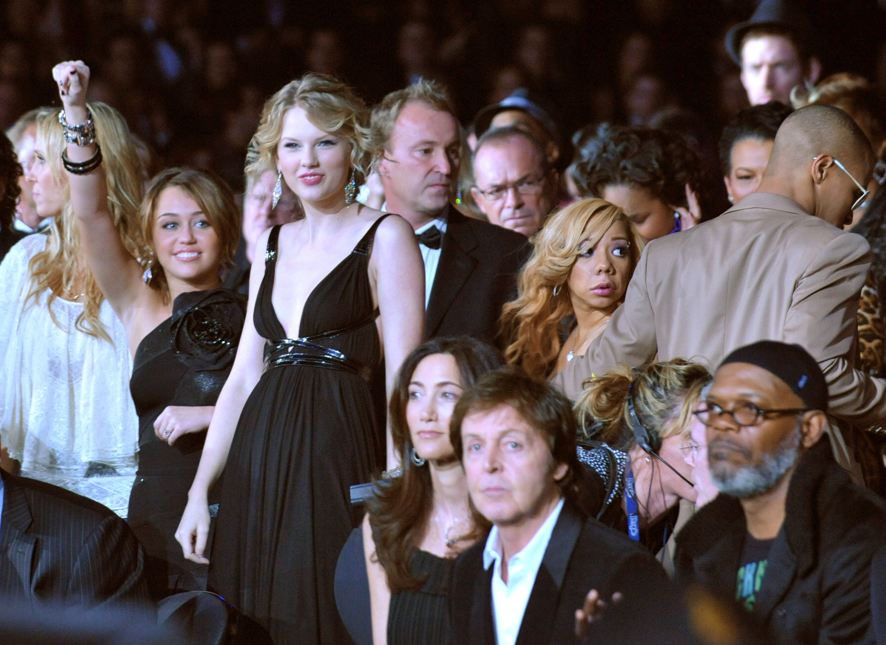 Miley Cyrus with a smile and her arm raised as she stands near Taylor Swift, Tiny, T.I., Paul McCartney, and Samuel L Jackson at an event