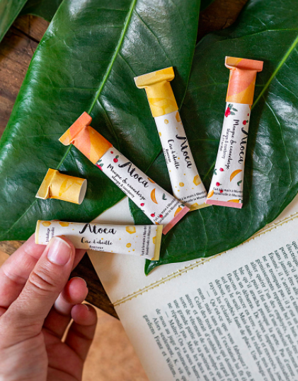A person holds up one of the lip balm tubes while the other three rest on a large monstera plant leaf