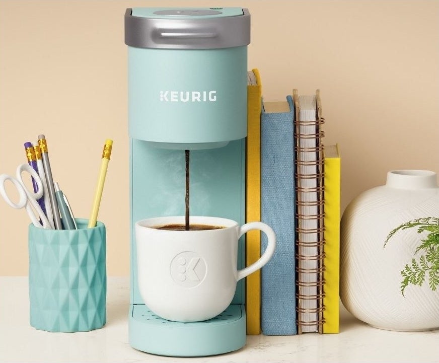 keurig k-mni single serve coffee maker sits on a decorated desk pouring coffee into a mug 