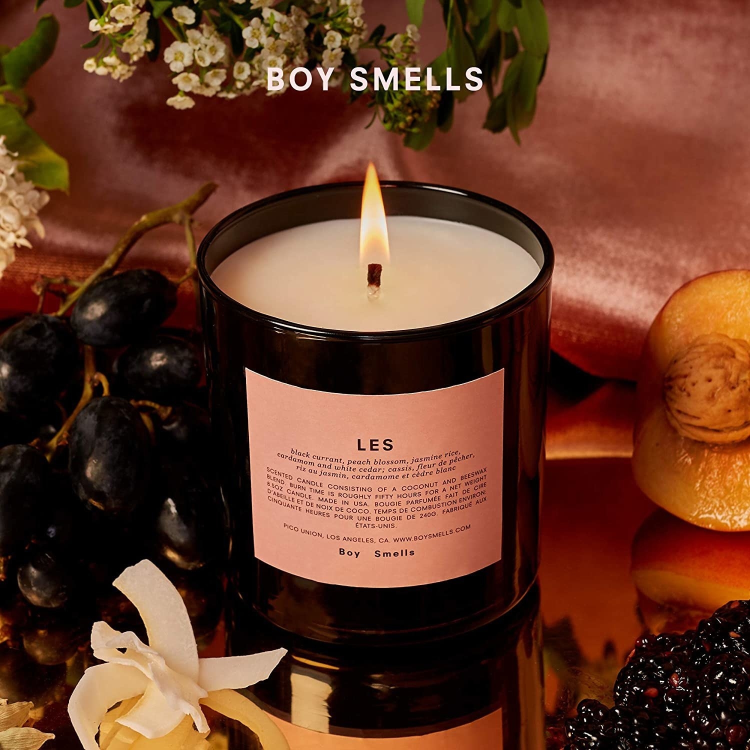 A lit LES candle on display with flowers, peaches, and grapes
