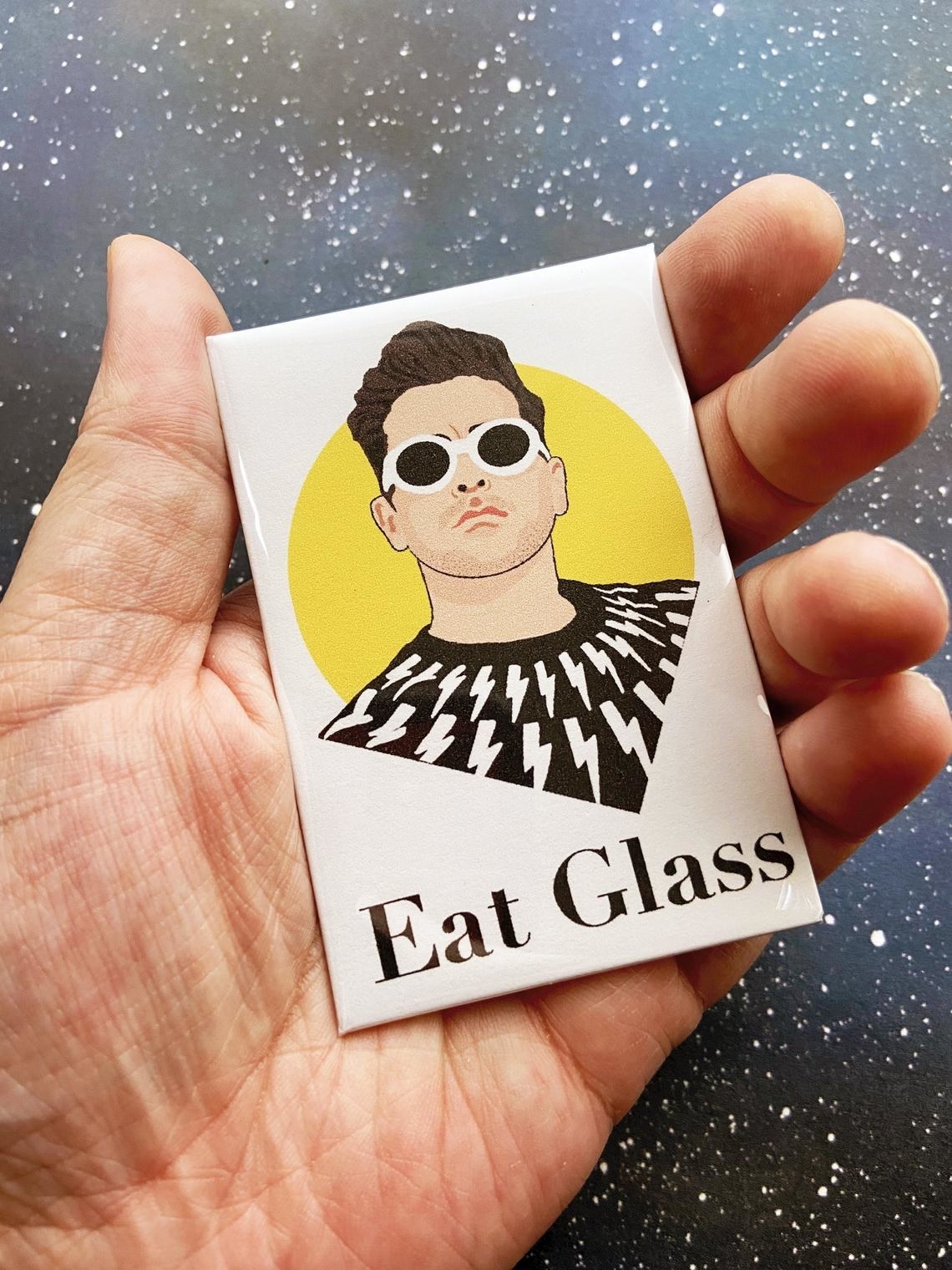 A hand holding a rectangular magnet featuring a portrait of David and the words &quot;Eat glass&quot;