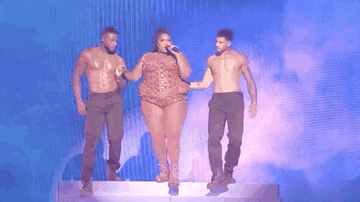 Lizzo walks down stairs in a leotard with two hunky men on her arms