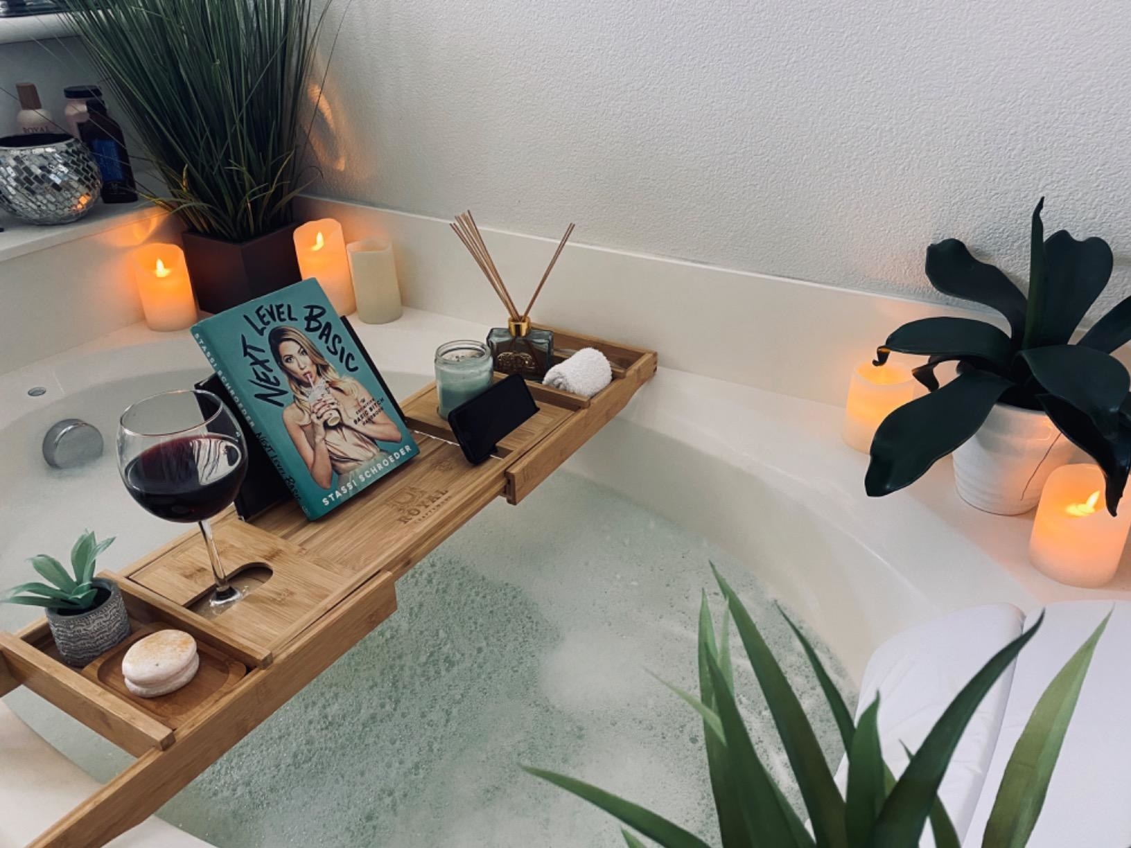 reviewer&#x27;s bath tub tray on the tub holding a book, wine, candle, and plants 