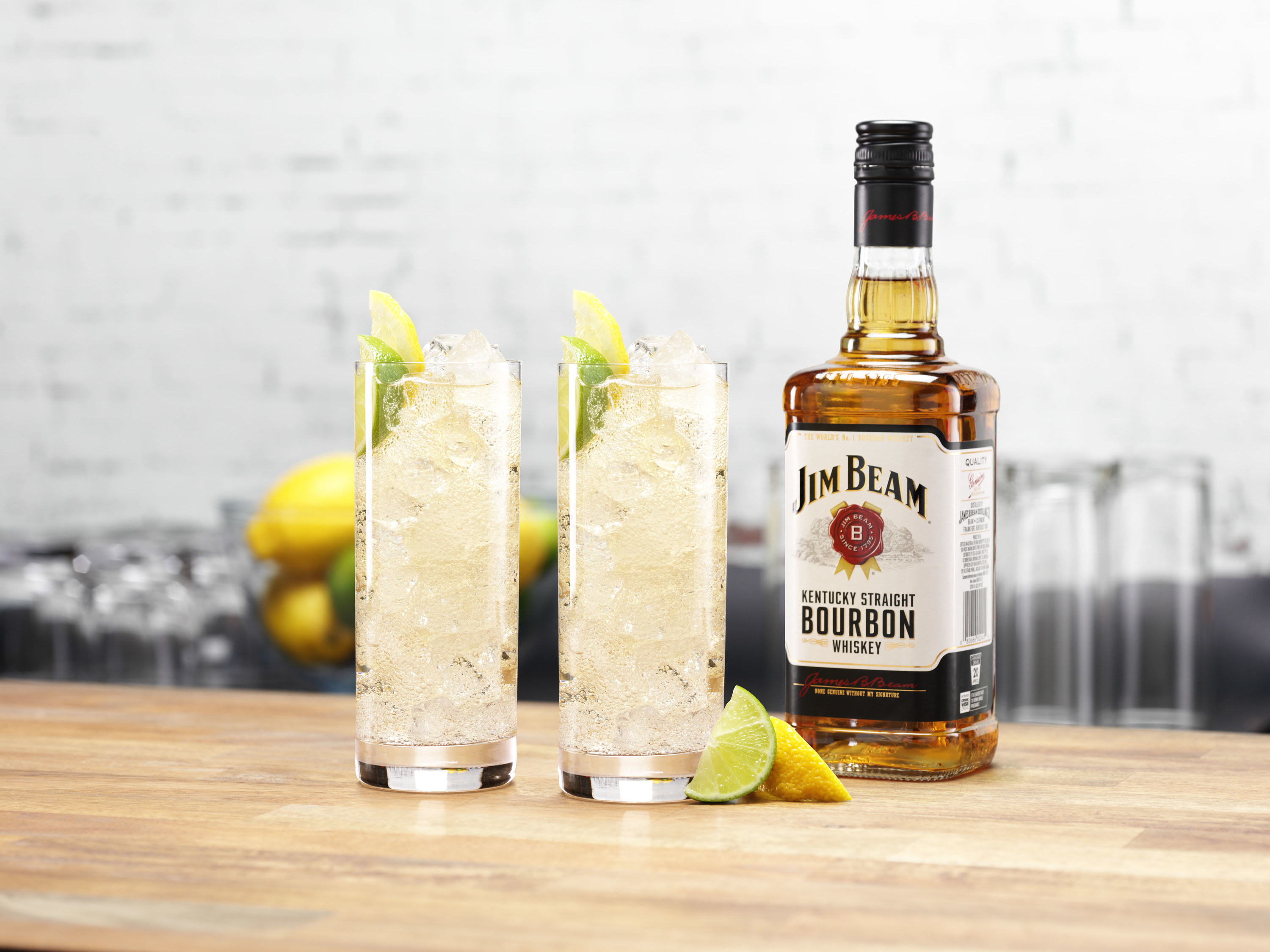 Two highball glasses filled with ice and liquid, garnished with lime and lemon next to a Jim Beam bottle 