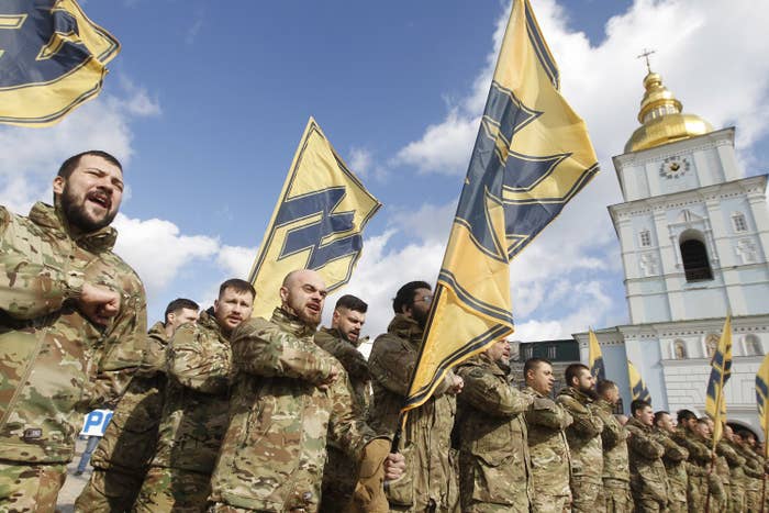 Men in military garb stand with flags featuring the Azov movement&#x27;s symbol, which is similar to the Wolfsangel, widely used by Nazi German divisions during the Second World War