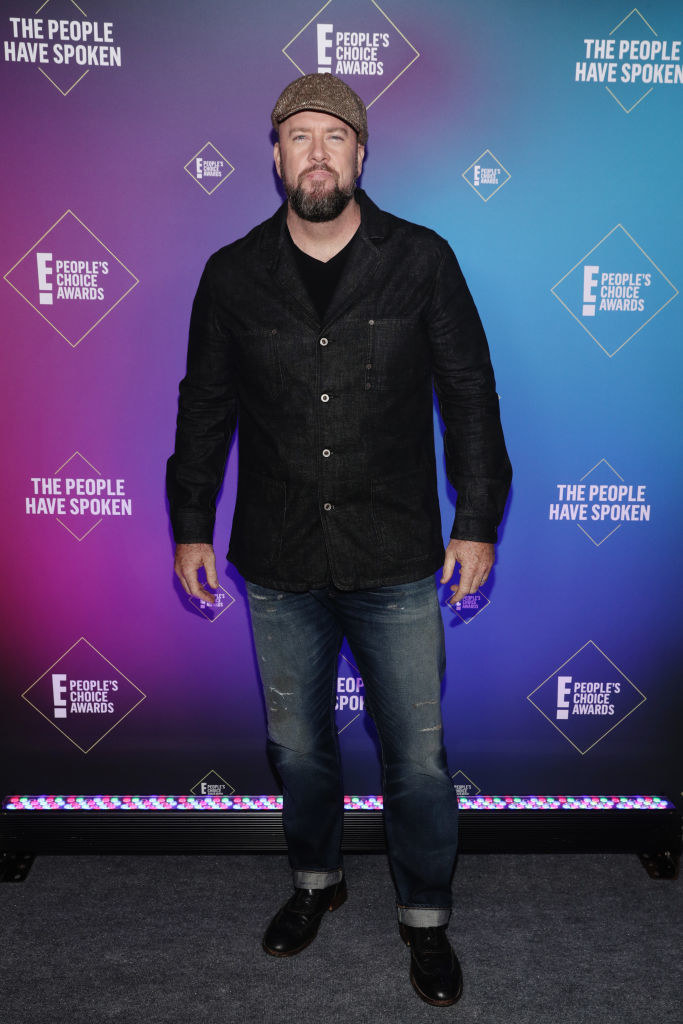 Chris Sullivan wears a button down shirt with distressed jeans and a textured newsboy cap