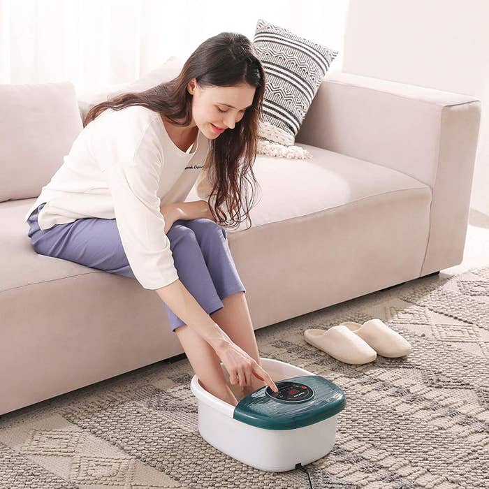Model presses button on top of green and white foot bath massager while sitting on a couch