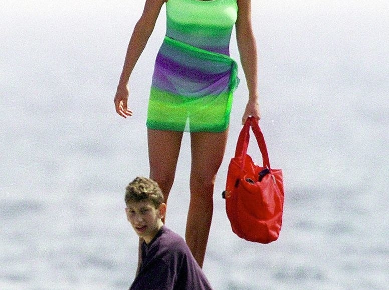 Princess Diana smiling in a bathing suit, a coverup tied around her waist, and sunglasses