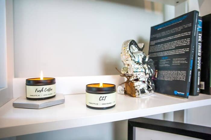 Tin soy candles that say &quot;Fresh Coffee&quot; and &quot;CLJ&quot; on a white shelf adorned with elephant bookends and novels