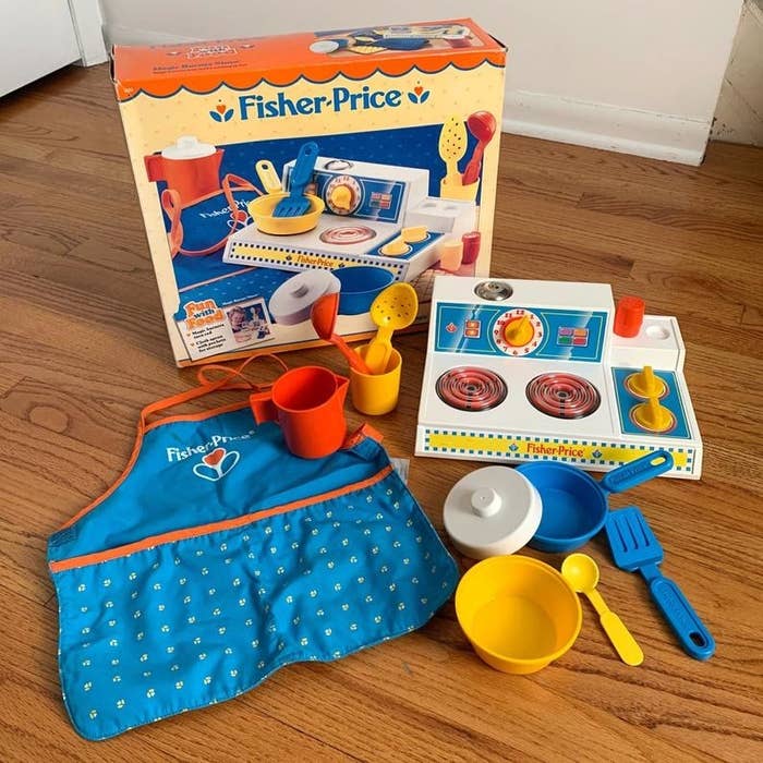 A Fisher-Price play cooking set featuring the box, a apron, a play stove top, and play cooking utensils and pots.