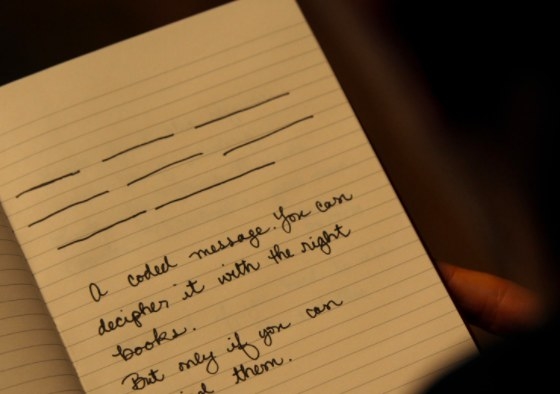 A close-up of an open notebook with blank lines drawn on the page and the words &quot;a coded message. you can decipher it with the right books.&quot; handwritten on it.