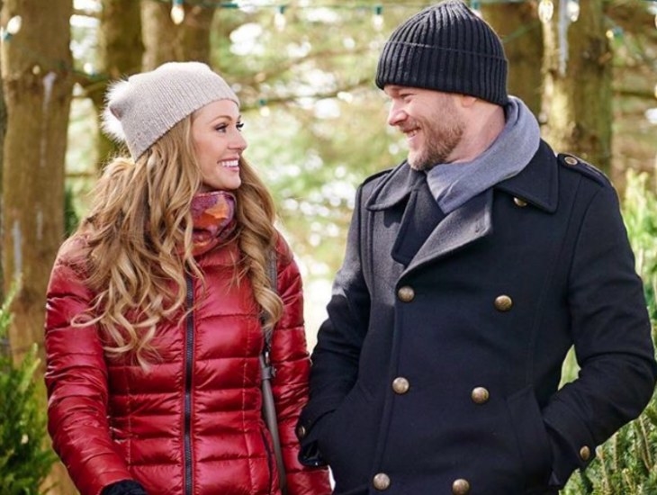 Rebecca Dalton and Aaron Ashmore, dressed in coats and beanies, stand in a sunlit forrest and smile at each other